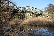 Un pont en métal dont les piles en briques sont taguées, au-dessus d’une petite rivière avec de hautes herbes.