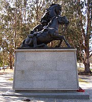A statue of two men and two horses stands atop a pedestal in a paved courtyard. Large trees provide a background.