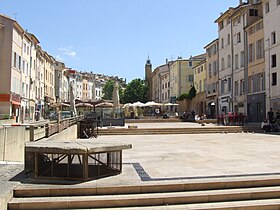 Photographie numérique d'une place ascendante sur trois plateaux et bordée de maisons. En arrière plan le sommet d'une tour horloge.