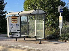 Un abribus vitré disposant à sa gauche d'une affiche publicitaire pour une chaîne de restauration rapide. À la gauche de l'image se trouve un poteau d'arrêt de bus où il est écrit "Saint-Cyr-sur-Menthon - Auberge".