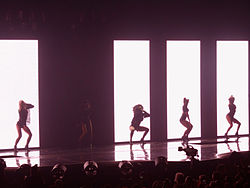 Una imagen que muestra a cinco mujeres vestidas con trajes negros frente a una pantalla