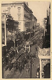 city street view from above, street seen diagonally from lower left to upper right, crowds of people line the street, horses, carriages and people march along it
