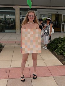 Femme portant une petite pancarte avec un patchwork de couleurs sur un ton rose en guise de robe. Portant seulement ce petit rectangle au niveau du buste, la personne semble nue.