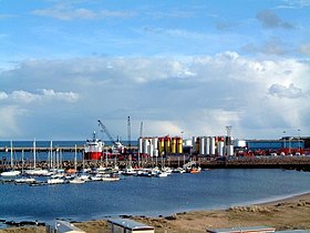 Peterhead Bay. In the background the breakwater built by convict labour. In the middle distance silos of drilling mud for the offshore oil industry and yachts berthed in the Peterhead marina. In the foreground the roofs of holiday caravans and the Lido sands.