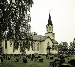 Fotografi av Rindal kirke med trevekst og kirkegård i forgrunnen.