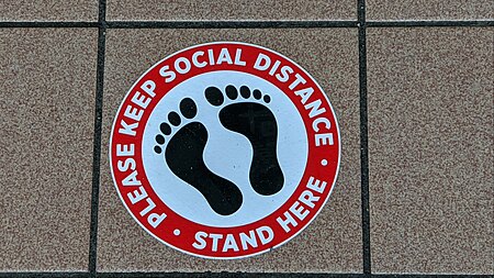 Circular sign with a sillhouette of bare feet, with text in a red border that says "Please keep social distance • Stand here"