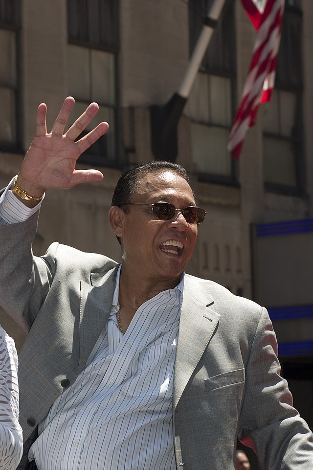 An olive-skinned man wearing sunglasses, a pinstriped shirt, and a gray jacket