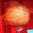12-panel brown leather football on a plinth of hardwood, with a brass panel inscribed with the names of the victorious team members, photographed through the glass of a display case