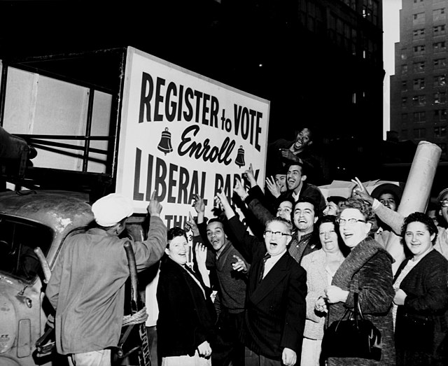 A cheering group of people point to a campaign banner that reads, "Register to Vote, Enroll Liberal Party."