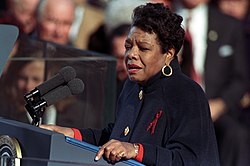 African American woman in her fifties, wearing a dark coat, standing at a lectern reading to a crowd gathered behind her.