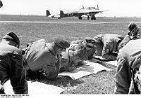 A group of men sitting and lying on a grass field reading maps, with a bomber aircraft in the background