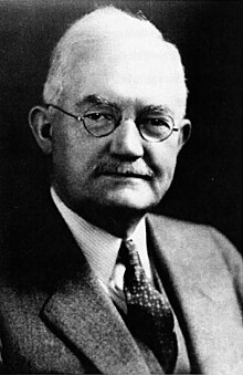 Black and white portrait photograph of an elderly man from the chest up. He wears a suit and glasses with round frames, and he has short, white hair.