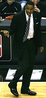 A man, wearing a black suit and white shirt, is standing on a basketball court while shouting and pointing at someone outside the picture.
