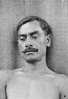 Photographie d'un homme torse nu regardant en bas à gauche et portant la moustache.