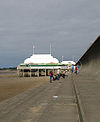 Kurzer Pier über Sand, überragt von einem weißen Pavillon mit Fahnenmasten.