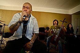 Homem com cabeça raspada, braços tatuados, cantando ao microfone na frente de músicos com instrumentos tradicionais