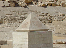 Pyramidion, který vypadá jako zmenšený model pyramidy, stojí na vyvýšeném kamenném stupínku. V pozadí se nachází zeď Červené pyramidy.