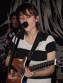 Sara Quin spielt mit einer Gitarre und singt in ein Mikrofon mit einem Schlagzeuger im Hintergrund