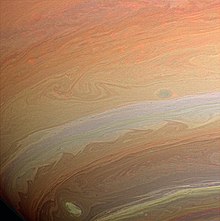 Fotografia de parte do planeta, em que aparecem faixas paralelas de coloração branca, alaranjada e vermelha com algumas ondulações e vórtices em suas bordas.