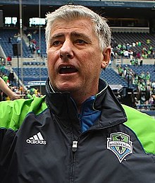 A man in a green and black tracksuit shaking hands with fans at a stadium.
