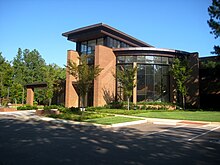 A tan brick building with dark glass walls, including a large curved glass wall facing the viewer. In front of the building are trees and a lawn, and in front of that a driveway and parking spaces.
