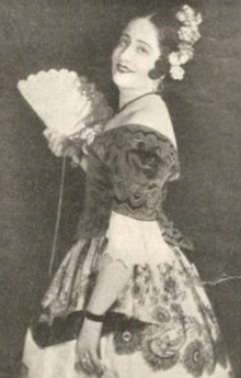 A smiling young white woman with dark hair, in an opera costume with bare shoulders, holding a fan