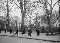 Млада жена се вози на ролшуи покрај група жени борци за право на глас пред Белата куќа, 1917