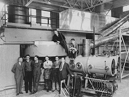 The 60-inch (1.52 m) cyclotron soon after completion in 1939. The key figures in its development and use are shown, standing, left to right: D. Cooksey, D. Corson, Lawrence, R. Thornton, J, Backus, W.S. Sainsbury. In the background are Luis Walter Alvarez and Edwin McMillan.