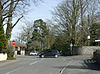 Street scene. Road junction with red stop sign. Two cars. The sides of the roads are stone walls with large trees showing above them.