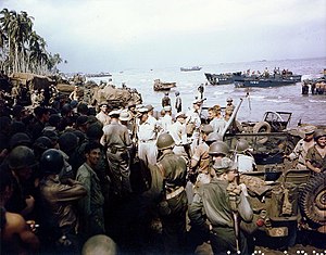 Kerumunan besar tentara dan jeep di pantai. Ada pohon kelapa di kejauhan dan landing craft lepas pantai. Kelompok kecil di tengah mencolok memakai seragam khaki dan memuncak topi bukan hutan hijau seragam dan helm.