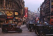 "A busy 1949 city scene in London, England, with black vintage vehicles, red London buses and many pedestrians. Buildings advertise "Wills's Gold Flake Cigarettes" and "Craven A" cigarettes.