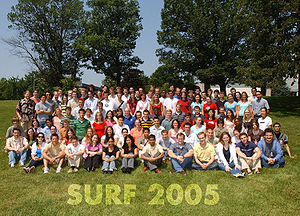 Color photo of a large number of students sitting on the grass with "Surf 2005" printed on the front.