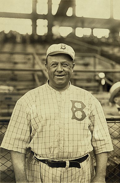 A man, wearing a baseball cap with a "B" in the center and a white baseball uniform with a square-pattern design and the Brooklyn Dodgers stylized "B" logo on the left breast, looks forward smiling.