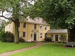 This tavern looks like a 2 1/2 story house, five windows wide and two deep. A single-story addition runs off one corner at an angle to the body of the house. The front door has a triangular pediment with dentil molding, and the appearance of rectangular pillars on either side of the door. An American flag hangs from a flagpole above the door.