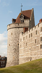 A photograph of a castle tower, the tower is pierced by small windows and has a coned, red-tiled roof, with a clock built into one side. The sky behind the wall is pale blue.