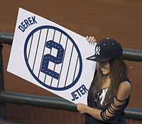 A woman with brown hair wearing a navy blue hat and navy blue shirt holds a sign to her right with the word "DEREK" at the top left, the word "JETER" at the bottom right, and a navy blue circle with navy blue vertical stripes and the number 2 inside it in the center.