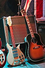 Two guitars lean against an amplifier. The left guitar is electric, and cream-coloured. The right guitar is acoustic, and brown and black in colour. A flag of Great Britain and a second amplifier are visible in the background, and part of a red electric guitar is visible to the left.
