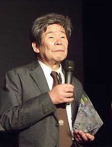 Mikrofona konuşan yaşlı bir adamın üç çeyrek boy portresi.