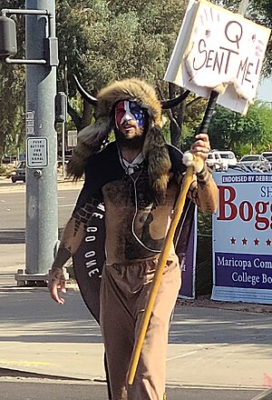 Angeli in his shaman dress on a sidewalk, holding a "Q sent me" sign