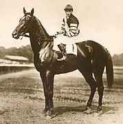 Photo en noir et blanc d'un jockey sur son cheval à l'arrêt, une piste de galop en arrière-plan.