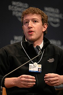 Waist high portrait of man in his thirties, looking into the camera and gesturing with both hands, wearing a black pullover shirt that says "The North Face" and wearing identification on a white band hanging from his neck