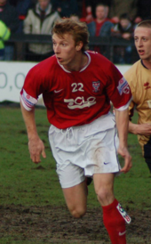 A man with brown hair is wearing a red top with a number and writing on the front, white shorts and red socks. He is standing on a grass field.