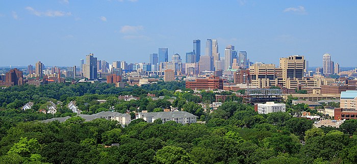 De skyline van Minneapolis stijgt naar het hoogste punt in het midden van het beeld, met de drie hoogste gebouwen die afsteken tegen een heldere blauwe lucht. Vóór de skyline zijn bomen, universiteitsgebouwen en wooncomplexen.
