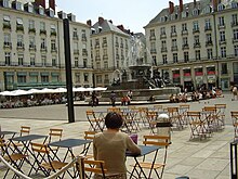 Des personnes sont attablées aux terrasses des cafés ou assise près de la fontaine dont l'eau jaillit.