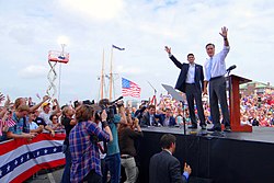 Mitt Romney and Paul Ryan seen in medium distance on an outdoor stage, with large crowd around them