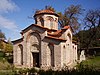 A mostly frontal view to a mid-sized medieval Byzantine-style church constructed out of bricks and white mortar. Greenery in the surrounding area.