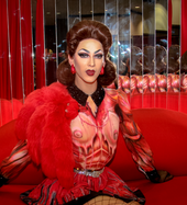 Porträtfoto von Violet Chachki, die auf einem roten Sofa sitzt. Sie trägt ein rotes Oberteil mit einem schwarzem, mit Edelsteinen besetzten Kragen und aufgemalten anatomischen Zeichnungen. Letzte stellen Sehnen, Blutgefäße und zwei Brüste dar. Daneben trägt sie eine Netzstrumpfhose, einen silbernen Gürtel, ein großes rotes Plüschstück an der rechten Körperseite und je einen runden, roten Ohrring an jedem Ohr. Außerdem trägt sie roten Lippenstift, aufgemalte Augenbrauen, Wimperntusche und rötlich-blauen Lidschatten. Ihre braunen Haare trägt als nach oben stehende, voluminöse, bis zum Hals reichende Frisur. Im Hintergrund sind mehrere längliche Spiegel zu sehen.