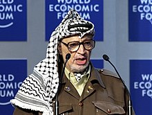 An elderly Palestinian man wearing a keffiyeh, a traditional Arab head  dress, and a pin with the portrait of late Palestinian leader Yasser Arafat  looks on