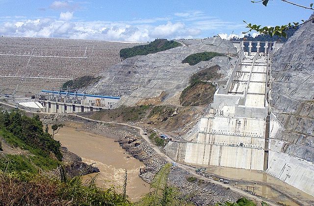 Image of a large rock structure and a power transformation station in the left, a rocky mountain, a river and another concrete structure in the right