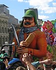 A parade float depicting a man with a thick mustache and a facial expression pinched in disgust, wearing a hunting cap, flannel shirt and scarf